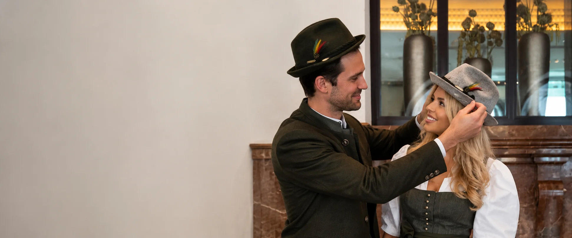 A man in a dark traditional outfit and bowler hat adjusts a gray hat on a woman wearing a white blouse and dark apron, set against an indoor background with a window and vases.